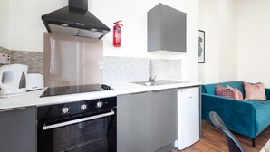 Une cuisine compacte équipée d’une cuisinière, d’un four et d’un lave-vaisselle, d’un meuble gris, d’un plan de travail blanc, d’un pare-feu gris, d’un extincteur rouge et d’un canapé turquoise dans l’espace salon.
