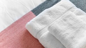 Une serviette blanche, soigneusement pliée, est posée sur un lit recouvert d’une couverture rose et grise.