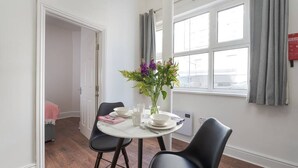 Un coin repas comprenant une table ronde, deux chaises, un vase avec des fleurs et une fenêtre avec des rideaux.
