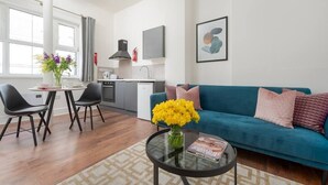 Un salon moderne avec un canapé turquoise, des chaises noires, une table basse ornée d’un vase de fleurs, et un coin cuisine en arrière-plan.