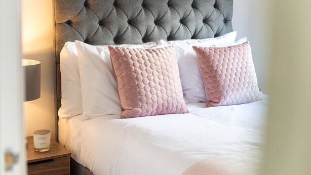 Un lit bien fait, avec une tête de lit capitonnée, des oreillers blancs et roses, et une table de chevet sur laquelle se trouvent une lampe et une bougie.