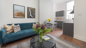 A modern living room with a teal sofa, a glass coffee table with a book, and a vase of flowers. There is a dining area with a white table and chairs, and a kitchen with a grey countertop and a TV mounted above it. - PBC | One Bed Apartment Earl's Court 121 EC2 (London)