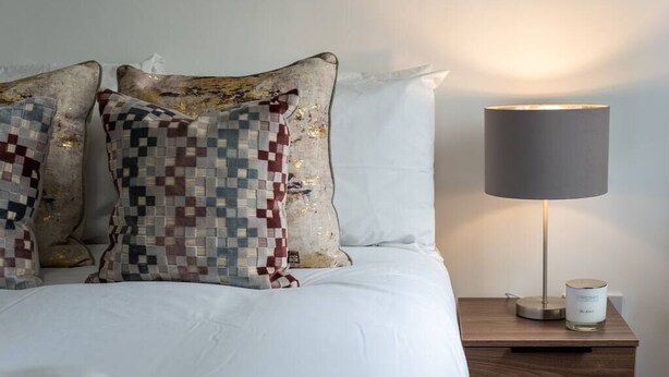 A neatly made bed with patterned pillows, a bedside lamp, and a small table with a candle. - PBC | One Bed Apartment Earl's Court 121 EC2 (London)