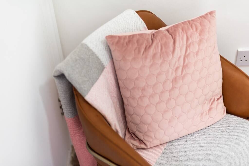 A chair with a grey and pink quilted pillow and a grey blanket.