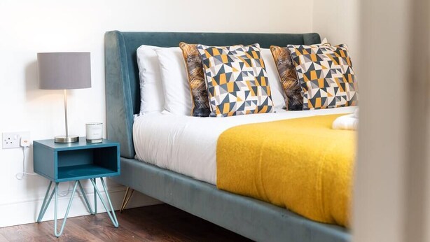 Une chambre à coucher bien rangée, avec un lit de couleur turquoise, une literie blanche et des coussins à motifs. Une petite table de chevet bleue, sur laquelle se trouvent une lampe et une bougie, est placée à côté du lit.