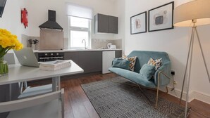 Une cuisine compacte dotée d’appareils électroménagers modernes, un coin repas avec un ordinateur portable et des livres, un canapé turquoise agrémenté de coussins et un tapis à motifs.
