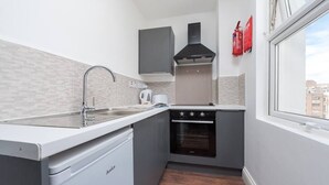 A compact kitchen with a dishwasher, oven, and a window with a view of buildings. - PBC | Studio Apartment Earl's Court 121 EC7 (London)