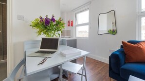 A modern living room with a white desk, a laptop, and a vase of flowers. There is a blue sofa with an orange cushion and a mirror on the wall. - PBC | Studio Apartment Earl's Court 121 EC7 (London)