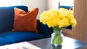A blue sofa with an orange pillow, a vase with yellow flowers, and a magazine on a table. - PBC | Studio Apartment Earl's Court 121 EC7 (London)