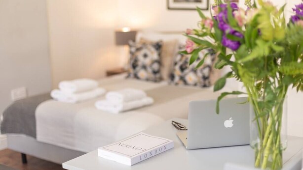 A neatly arranged hotel room with a bed, a laptop, a book titled "London," and a vase with flowers. - PBC | Studio Apartment Earl's Court 121 EC7 (London)