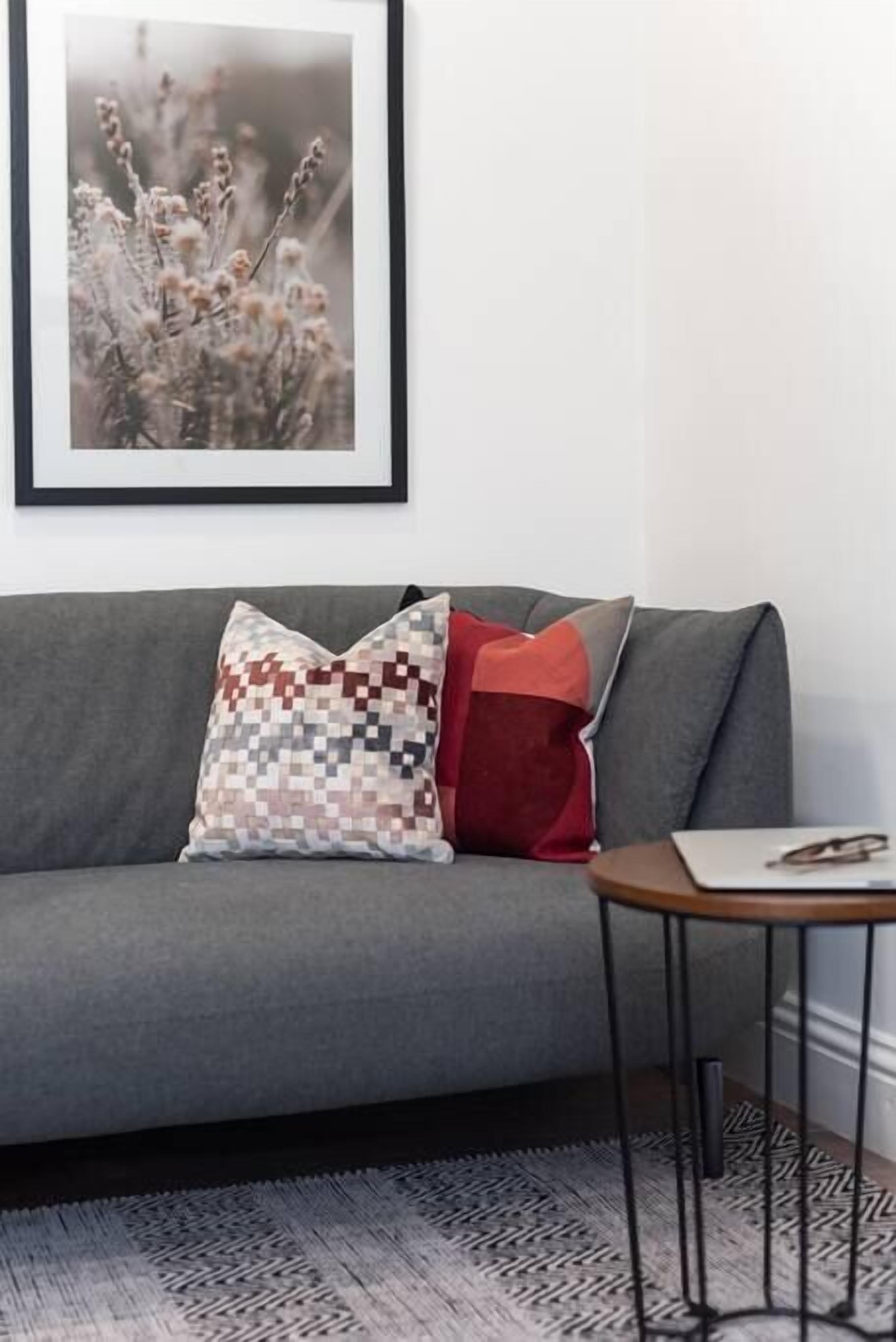 A grey sofa with a framed botanical print, a pixelated cushion, and a red pillow. A small wooden side table with a laptop and glasses is beside the sofa.