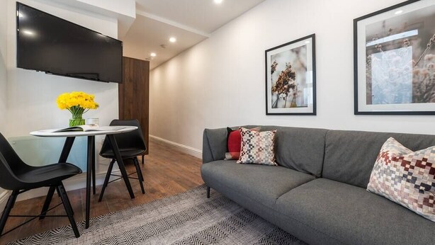 A modern living room with a grey sofa, black chairs, a round table with a vase of yellow flowers, and two framed pictures on the wall. - PBC | Studio Apartment Earl's Court 119C ECA1 (London)