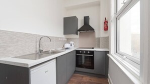 Une cuisine compacte avec des meubles gris, un plan de travail blanc, un lave-vaisselle Bosch, un four noir et une fenêtre qui laisse entrer la lumière naturelle.