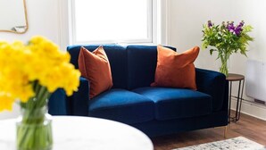 Un canapé bleu avec des coussins orange, une table ronde blanche ornée d’un vase de fleurs jaunes et une petite table d’appoint avec un vase de fleurs violettes.