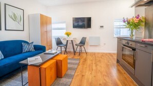 Eine moderne Wohnung mit einem blauen Sofa, einem kleinen Esstisch mit Stühlen, einer Küche mit Herd und einem Fernseher an der Wand.