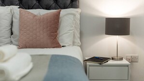A neatly made bed with white and pink pillows, a bedside table with a lamp, and a notebook. - PBC | One Bed Earl's Court ECC3 (London)
