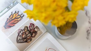 A book with abstract art on the pages, a vase with yellow flowers, and a metal object on the table. - PBC | One Bed Earl's Court ECC3 (London)