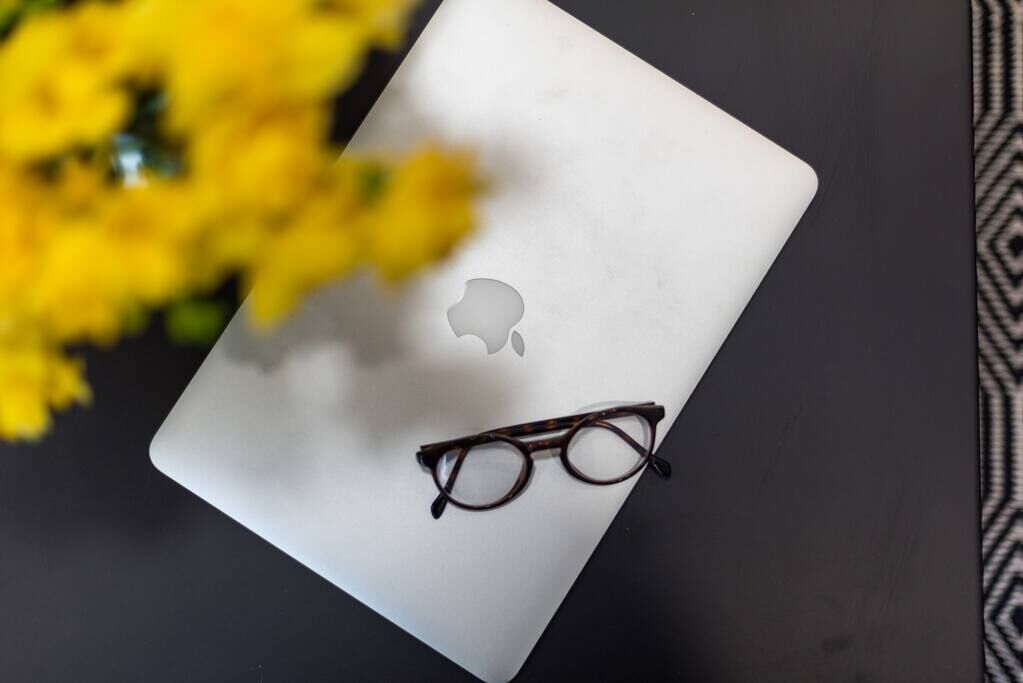 A laptop with a pair of glasses on top, placed on a dark surface with yellow flowers in the background.