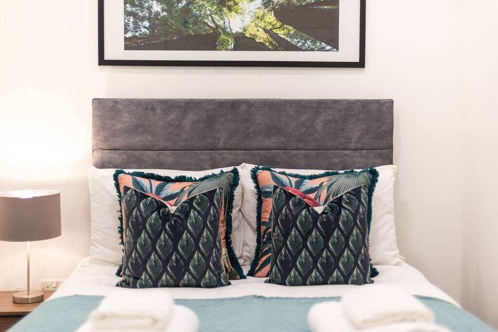 Ein ordentlich bezogenes Bett mit grauem Kopfteil, zwei Zierkissen, einem gerahmten Bild darüber und einer Nachttischlampe.