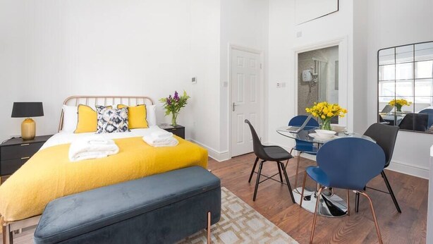 A modern bedroom with a bed, bedside table, and a small desk area with chairs and a vase of flowers.