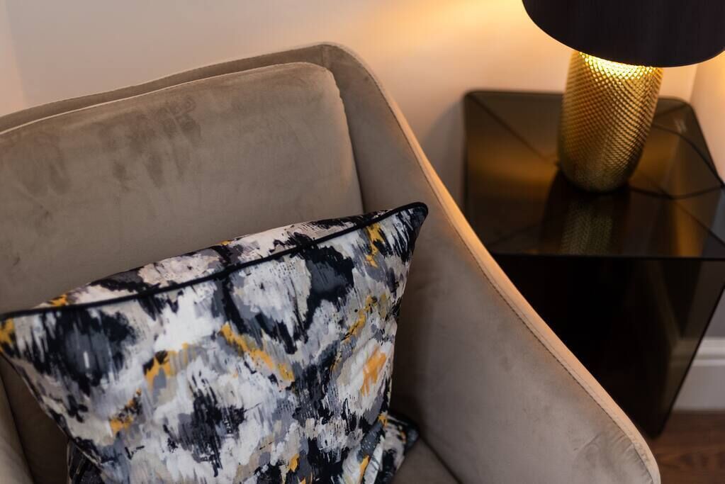 A modern armchair with a textured cushion, a black and gold lamp on a black side table, and a neutral wall in the background.