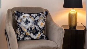 Een beige fauteuil met een bloemenkussen, een zwart metalen frame en een gouden lamp op een klein zwart bijzettafeltje.