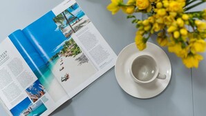 An open magazine with travel photos, a white cup on a saucer, and a vase of yellow flowers on a table. - PBC | Cosy 1-Bed Apt - Earl's Court 119 - EC3 (London)