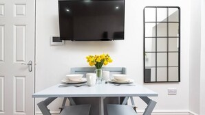 Een eetgedeelte met een witte tafel, grijze stoelen, een flatscreen televisie aan de muur en een spiegel.