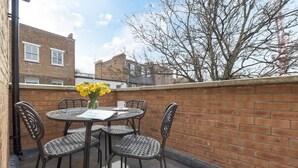 Een terras met een ronde tafel en vier rieten stoelen, een vaas met gele bloemen, en een bakstenen muur.
