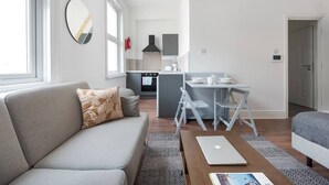 A modern living room with a grey sofa, a coffee table, and a dining area with a small table and chairs. - PBC | Large Studio - Earl's Court 119 - EC6 (London)