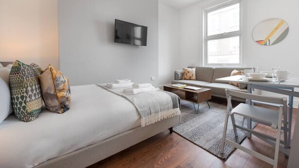 A modern, well-lit room with a bed, a sofa, a small table, and a TV mounted on the wall. - PBC | Large Studio - Earl's Court 119 - EC6 (London)