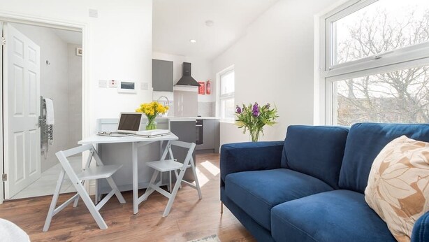 A modern living room with a blue sofa, a dining table with chairs, and a kitchen area in the background. - PBC | Studio Apartment Earl's Court 119 EC7 (London)