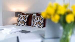 A neatly made bed with patterned pillows, a bedside lamp, and a vase with yellow flowers. - PBC | Studio Apartment Earl's Court 119 EC7 (London)