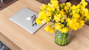 Een laptop met een bril op een houten tafel, samen met een vaas gele bloemen.