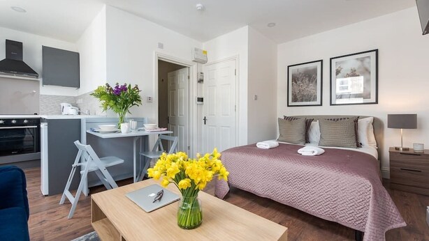 Een compacte hotelkamer met een bed, nachtkastje en een klein bureau met stoel. Achtergrond toont een kitchenette.
