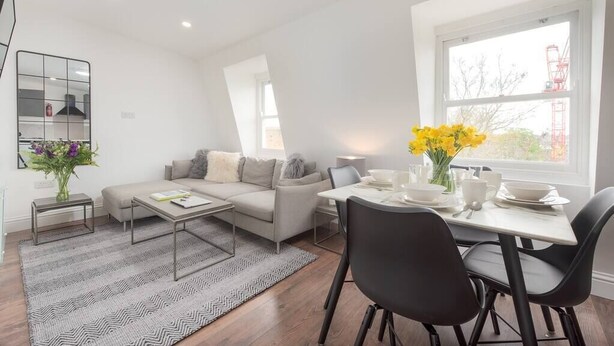 A modern living room with a sofa, coffee table, and dining area with chairs and a table set for two. - PBC | Two-Bed Apt - Earl's Court 119 - EC9 (London)