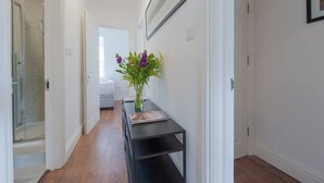 A hallway with a wooden floor, a black console table with a vase of flowers, and a framed picture on the wall. - PBC | Two-Bed Apt - Earl's Court 119 - EC9 (London)