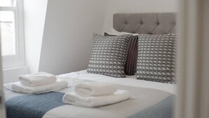 A neatly made bed with a tufted headboard, two pillows with geometric patterns, and folded towels on the bed. - PBC | Two-Bed Apt - Earl's Court 119 - EC9 (London)