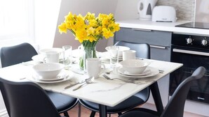A dining table set with white plates, bowls, and cutlery, accompanied by a vase of yellow flowers. - PBC | Two-Bed Apt - Earl's Court 119 - EC9 (London)