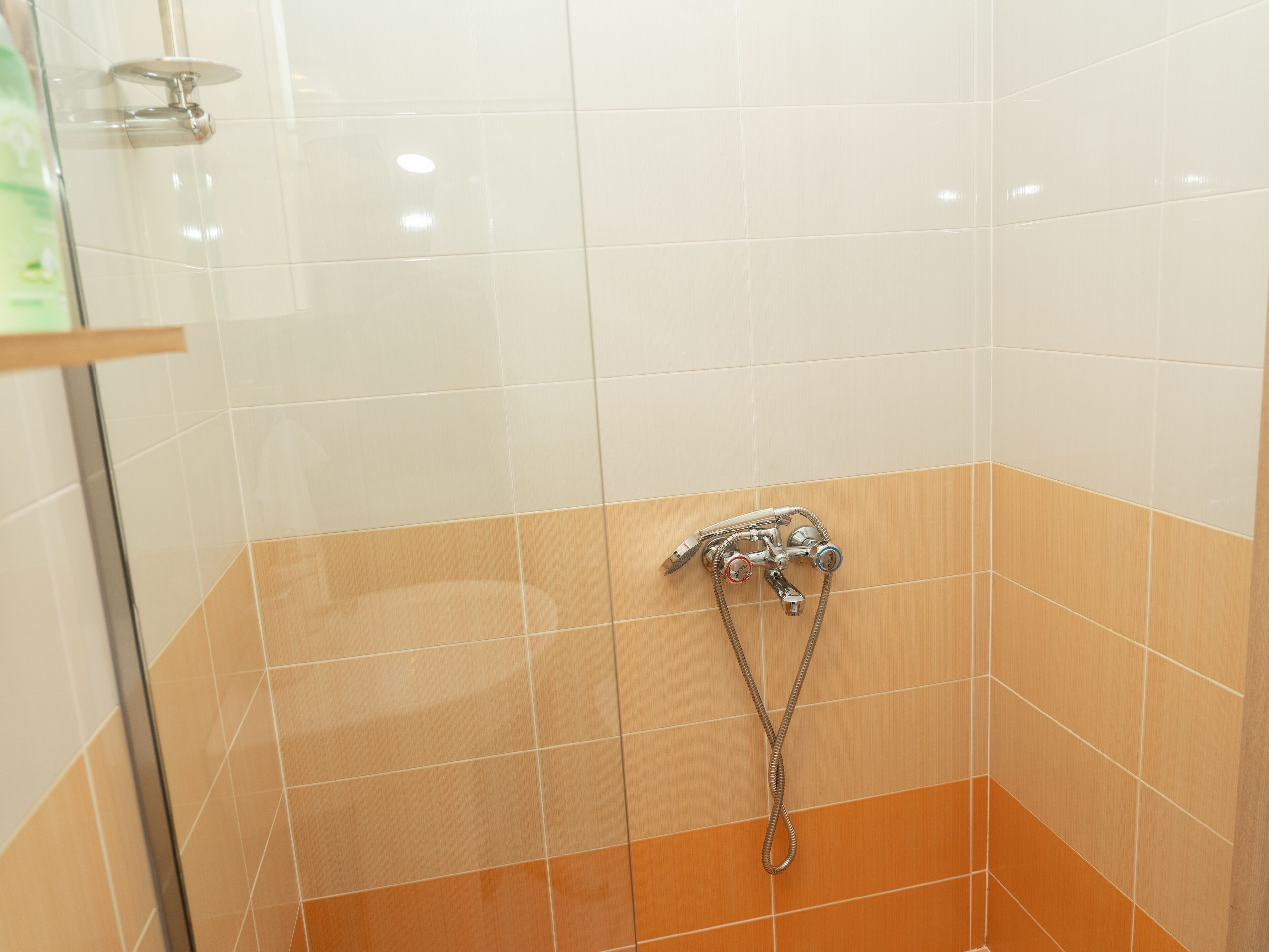 Un área de ducha con azulejos blancos y naranja, una puerta de cristal y un cabezal de ducha con múltiples boquillas.