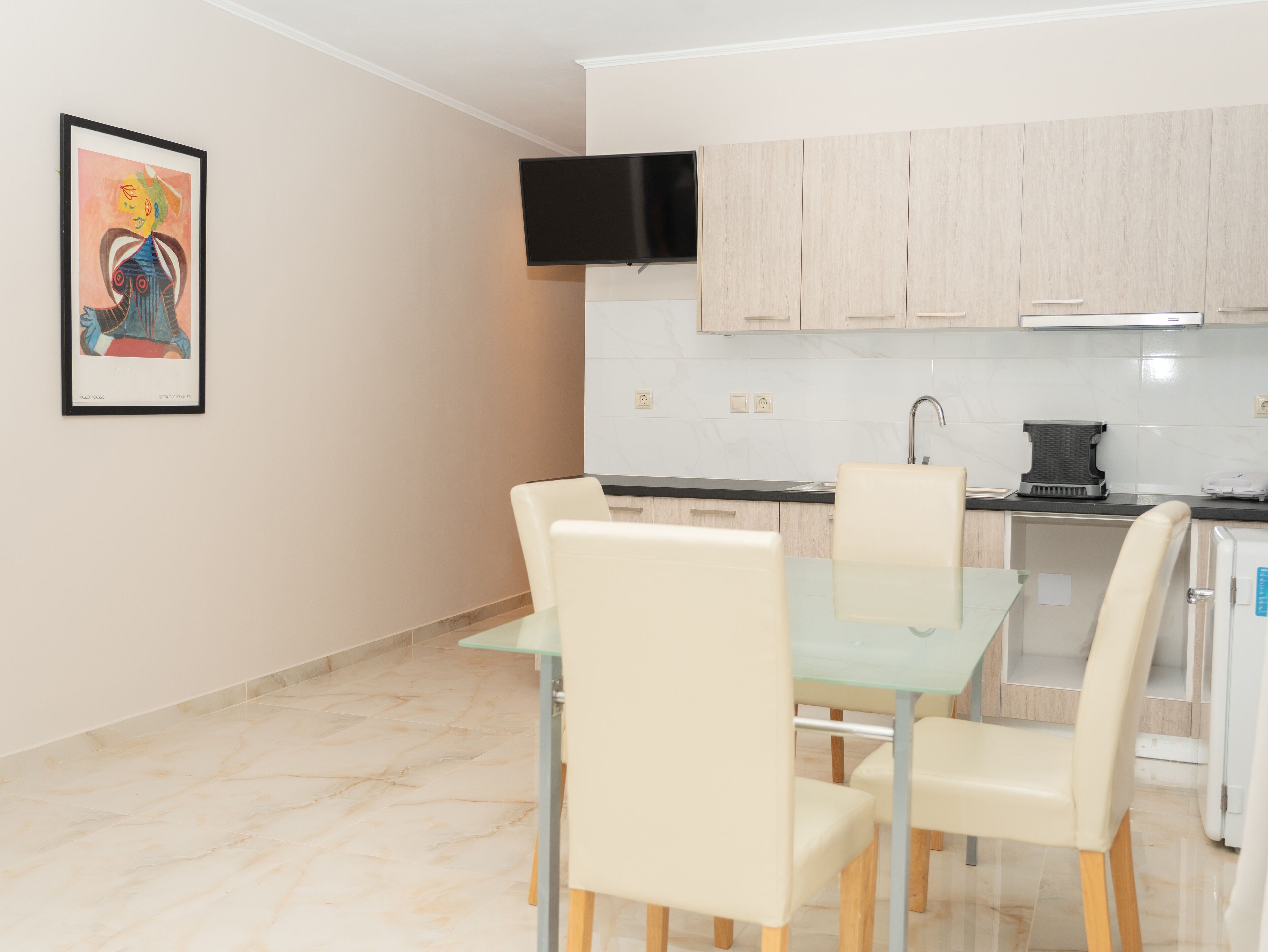 A modern kitchen with light wood cabinets, a black TV mounted above the counter, and a glass dining table with four chairs.