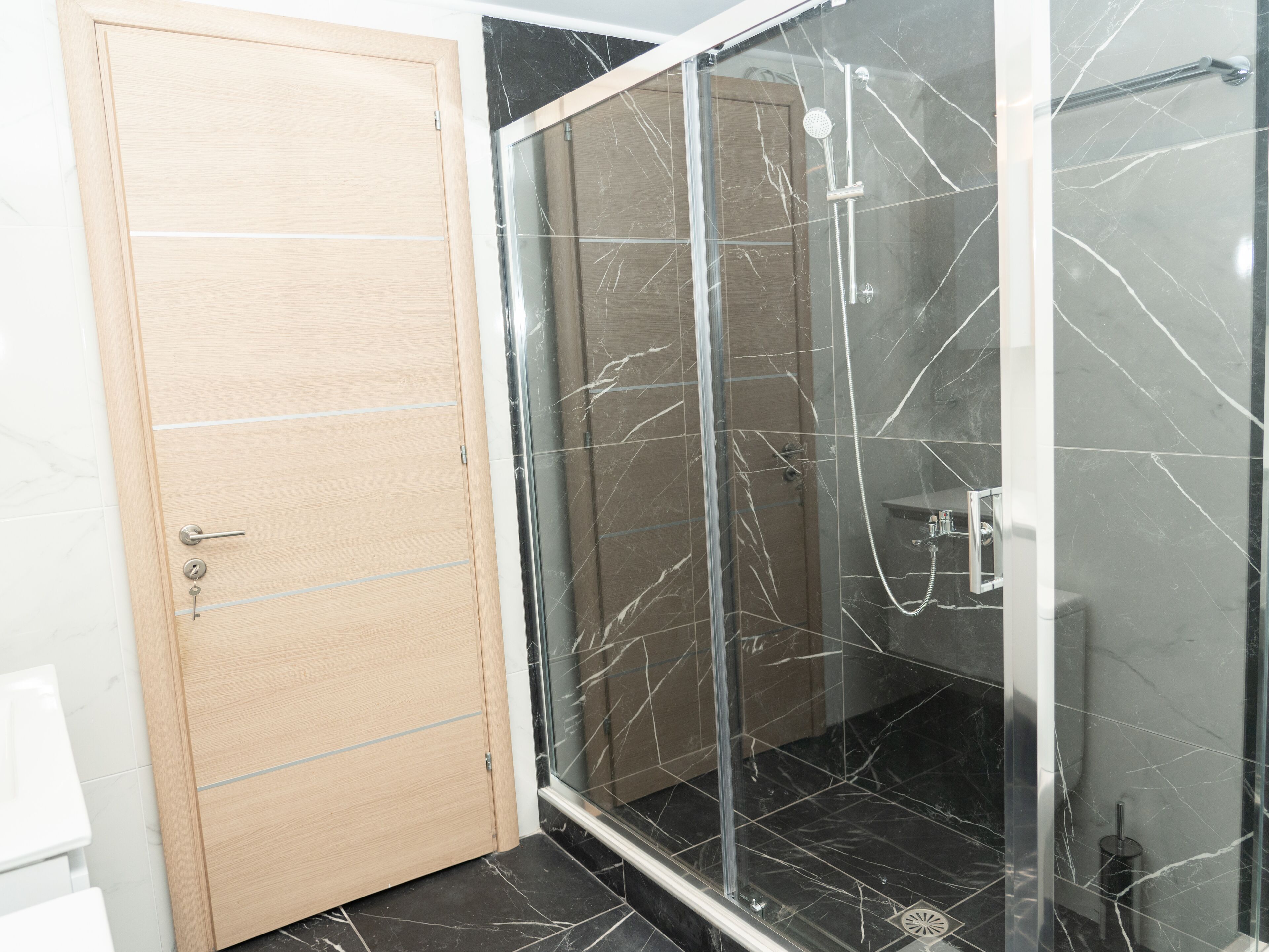 A modern bathroom with a wooden door, a glass shower enclosure, and black marble tiles.