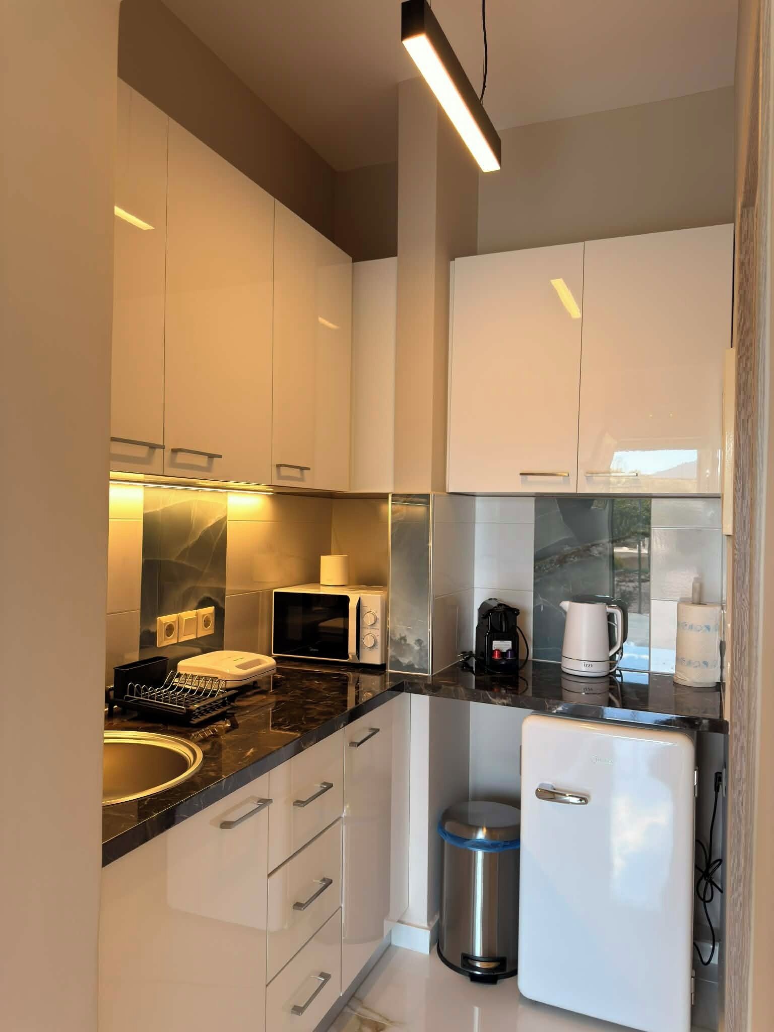 A modern kitchen with white cabinets, a black countertop, a microwave, a coffee maker, and a trash bin.