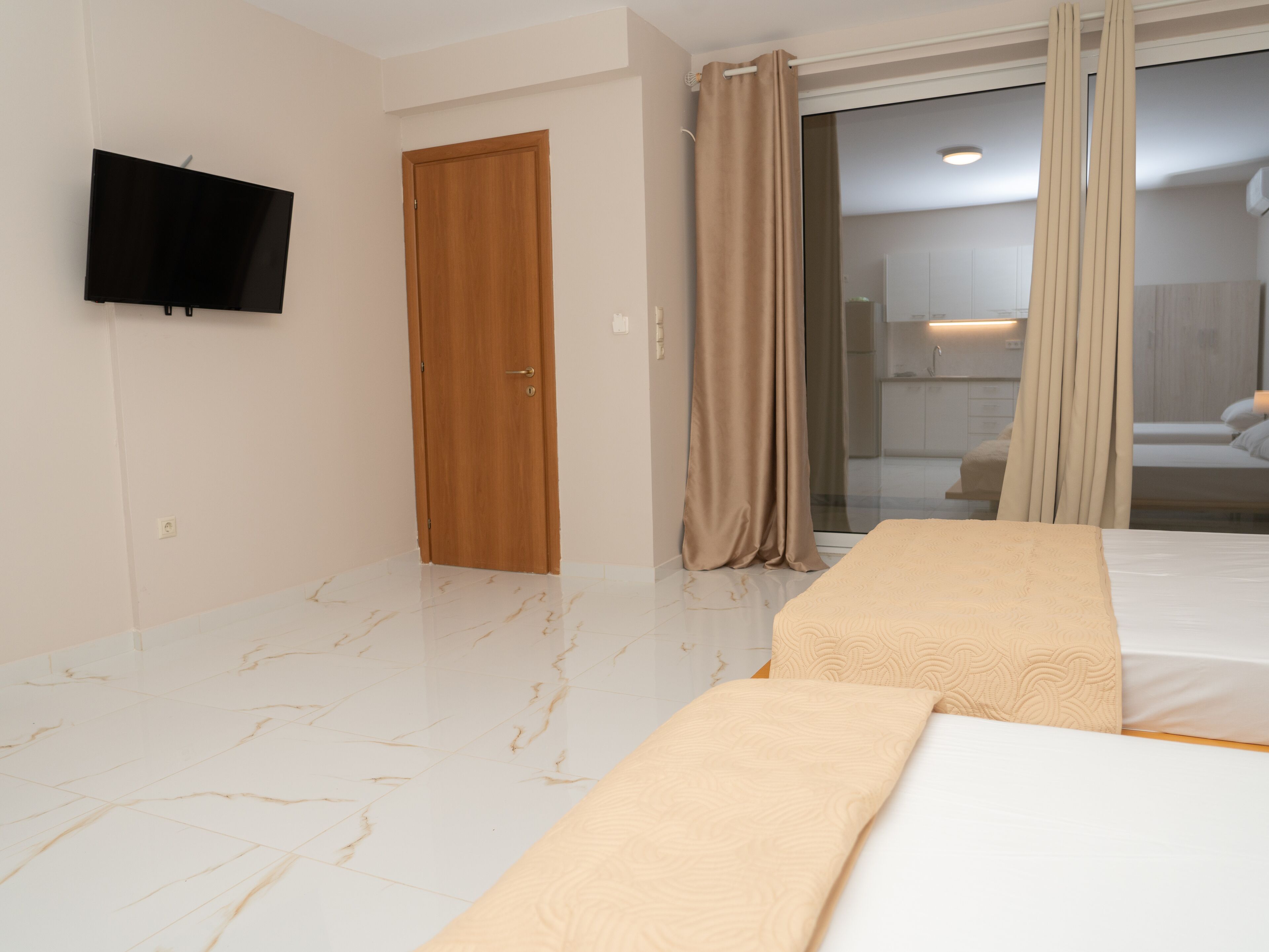 Una habitación de hotel moderna con televisión de pantalla plana montada en la pared, una puerta de madera, una puerta corredera de vidrio que da al baño, y dos camas con ropa de cama color beige.