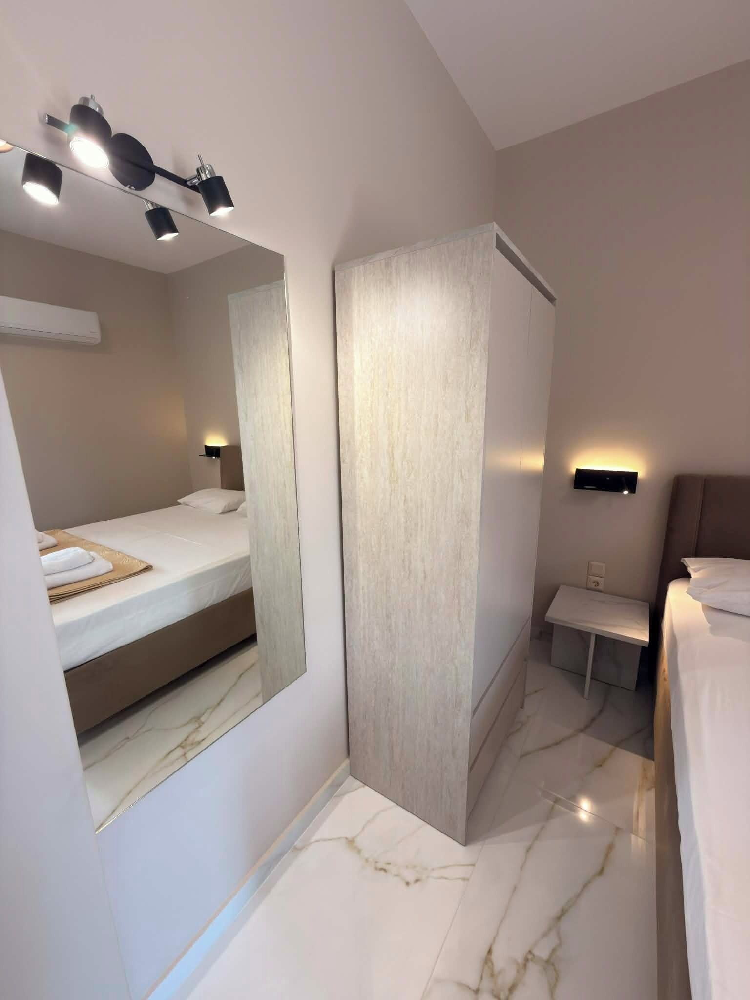 A modern hotel room with a large mirror, a bed, a small bedside table, and a wall-mounted air conditioning unit.