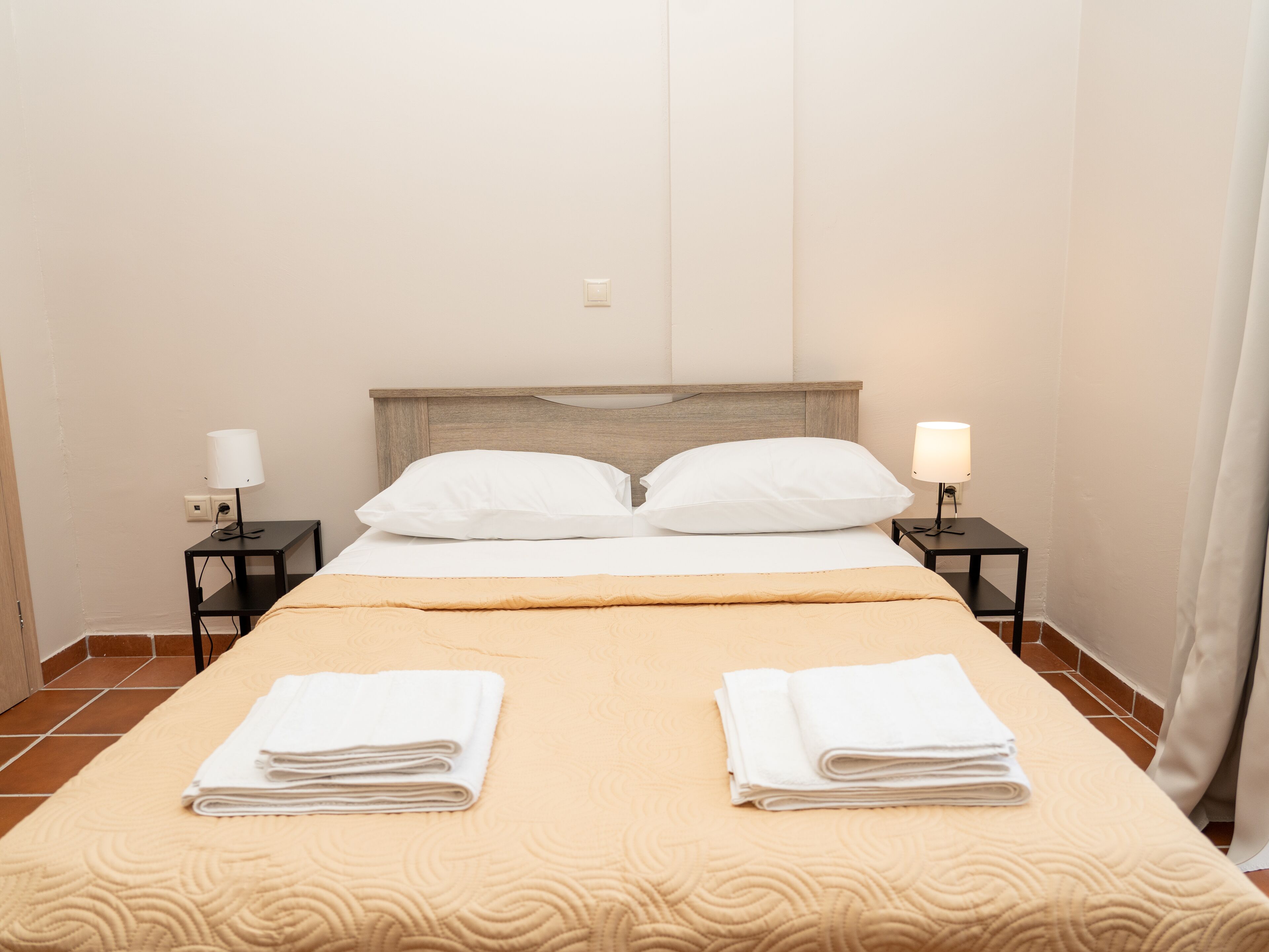 A neatly made bed with two pillows, a beige bedspread, and two folded white towels on the bed. There are two bedside tables with lamps and a power outlet on the wall.