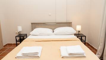 A neatly made bed with two pillows, a beige bedspread, and two folded white towels on the bed. There are two bedside tables with lamps and a power outlet on the wall.
