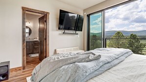 A bedroom with a large bed, a TV mounted on the wall, and a balcony with a view of the countryside. - New Luxury w/ AC | Close to Slopes | Hot Tub | Valley View Villa 11 (Angel Fire)