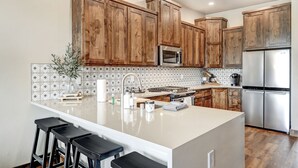 A kitchen with wooden cabinets, stainless steel appliances, and a central island with stools. - New Luxury w/ AC | Close to Slopes | Hot Tub | Valley View Villa 11 (Angel Fire)