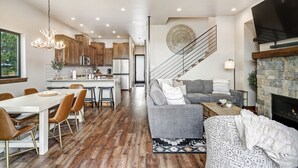 A modern kitchen with wooden cabinets, a dining area with a white table and brown chairs, a gray sofa, and a fireplace. - New Luxury w/ AC | Close to Slopes | Hot Tub | Valley View Villa 11 (Angel Fire)